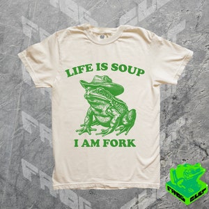 May include: A light beige t-shirt with a green frog wearing a cowboy hat. The text "Life is Soup I am Fork" is printed on the shirt.