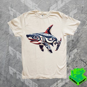 May include: A white t-shirt with a blue, red, and white stylized illustration of a shark. The shark is depicted in a Native American art style. The text "FROG CULT" is printed on the bottom right corner of the shirt.