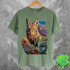 May include: A green t-shirt with a colorful graphic print of a coyote in a desert landscape. The coyote is looking to the left, and the landscape includes cacti and mountains.