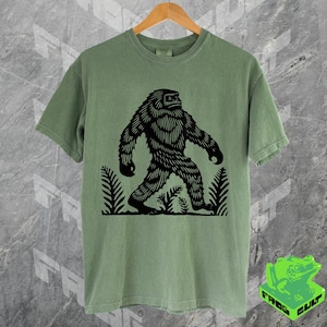 May include: A green t-shirt with a black silhouette of Bigfoot walking through a forest. The t-shirt is made of a soft, comfortable material.