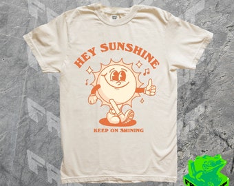 May include: A beige t-shirt with an orange cartoon sun graphic. The sun is smiling and giving a thumbs up. The text "Hey Sunshine Keep on Shining" is printed on the shirt.