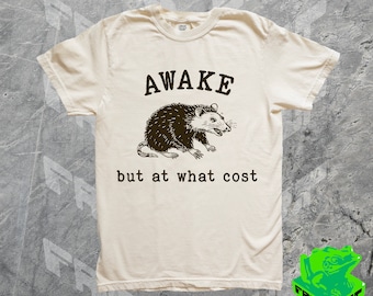 Pode incluir: Uma camiseta bege com uma ilustração em preto e branco de um gambá. O texto "AWAKE but at what cost" está impresso na camiseta.