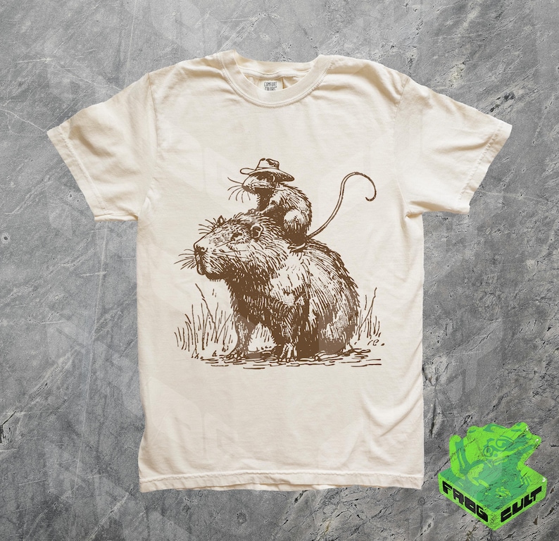 Puede incluir: Una camiseta beige con una ilustraci&oacute;n marr&oacute;n de una capibara con un sombrero de vaquero y montando un rat&oacute;n. El rat&oacute;n tambi&eacute;n lleva un sombrero de vaquero.