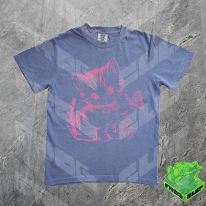 May include: A blue t-shirt featuring a pink graphic of a kitten with a butterfly. The kitten is sitting and has a bow around its neck. The shirt has short sleeves and a crew neck.