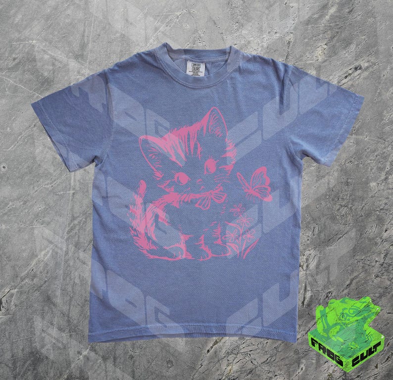 May include: A blue t-shirt featuring a pink graphic of a kitten with a butterfly. The kitten is sitting and has a bow around its neck. The shirt has short sleeves and a crew neck.
