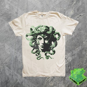 May include: A white t-shirt featuring a green graphic print of Medusa, a mythical Greek figure with snakes for hair.