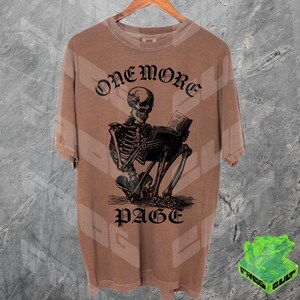 May include: Brown t-shirt with a graphic of a skeleton reading a book. The text "ONEMORE PAGE" is above and below the skeleton. The shirt is hanging on a wooden hanger.