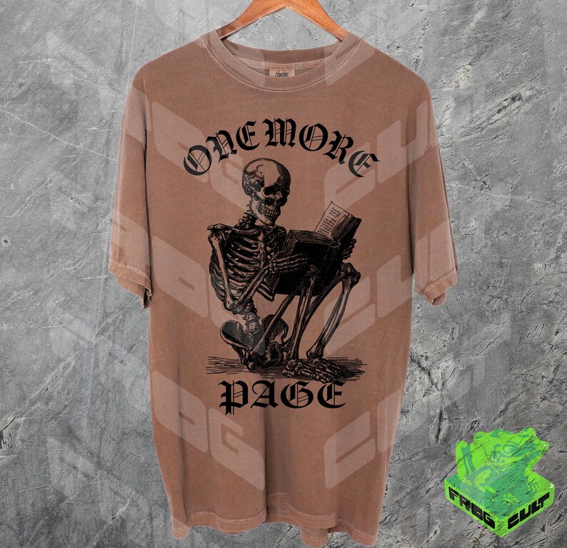 May include: Brown t-shirt with a graphic of a skeleton reading a book. The text "ONEMORE PAGE" is above and below the skeleton. The shirt is hanging on a wooden hanger.