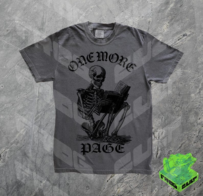 May include: Gray t-shirt featuring a black illustration of a skeleton reading a book. The text "ONEMORE" is above the skeleton, and "PAGE" is below. The shirt has a crew neck and short sleeves.