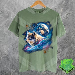 Puede incluir: Una camiseta verde con un lobo aullando a la luna en un estampado de galaxia. El lobo es gris y blanco con ojos azules. La luna es una luna creciente con una galaxia azul y blanca detr&aacute;s.