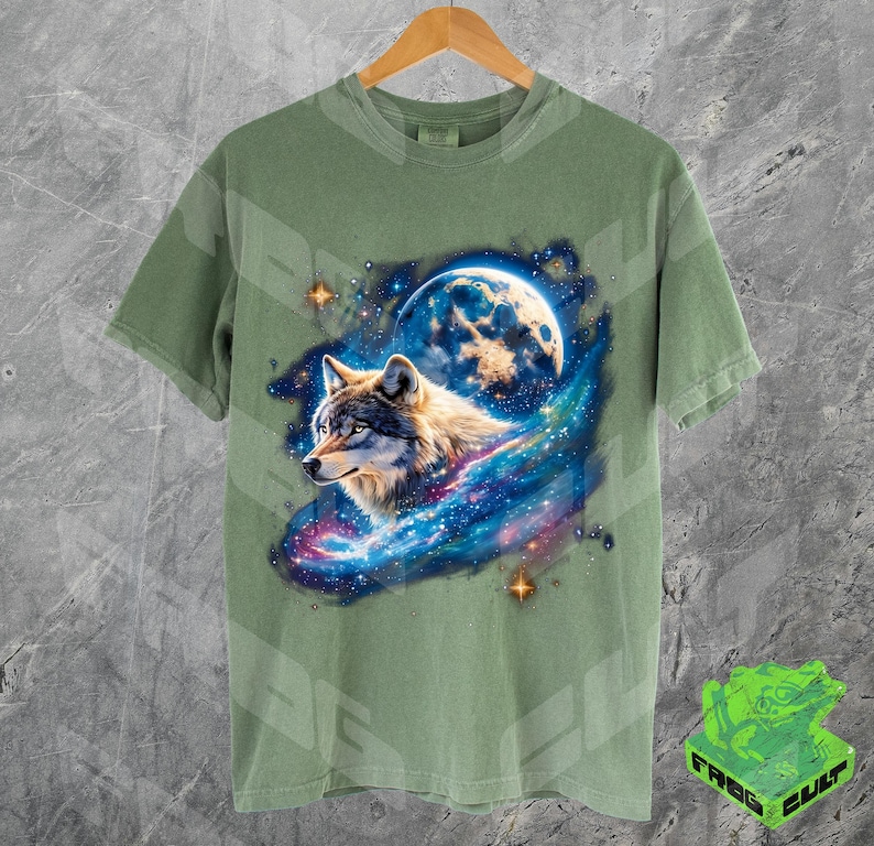 Puede incluir: Una camiseta verde con un lobo aullando a la luna en un estampado de galaxia. El lobo es gris y blanco con ojos azules. La luna es una luna creciente con una galaxia azul y blanca detr&aacute;s.