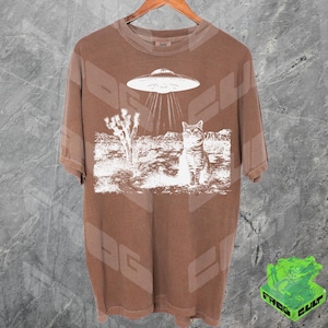 Könnte beinhalten: Ein braunes T-Shirt mit einem weißen Aufdruck eines Katers, der zu einem UFO in einer Wüstenlandschaft aufblickt. Der Text "Alien Cat" ist auf dem Shirt gedruckt.