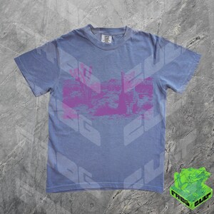 May include: A light blue t-shirt with a pink graphic print of a desert landscape. The shirt has a crew neck and short sleeves.