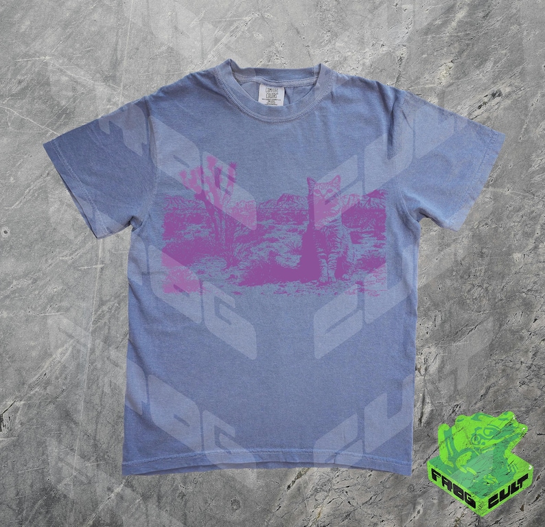 May include: A light blue t-shirt with a pink graphic print of a desert landscape. The shirt has a crew neck and short sleeves.