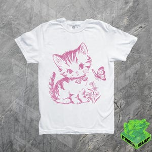 May include: White t-shirt featuring a pink illustration of a kitten with a bow, flowers, and a butterfly. The graphic is centered on the front of the shirt. The shirt has short sleeves and a crew neck.