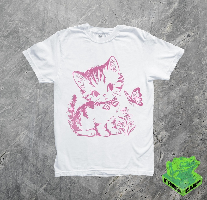 May include: White t-shirt featuring a pink illustration of a kitten with a bow, flowers, and a butterfly. The graphic is centered on the front of the shirt. The shirt has short sleeves and a crew neck.