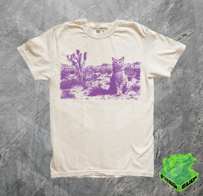 May include: A white t-shirt with a purple print of a cat sitting in a desert landscape. The cat is looking to the left. The print includes a Joshua tree in the background.