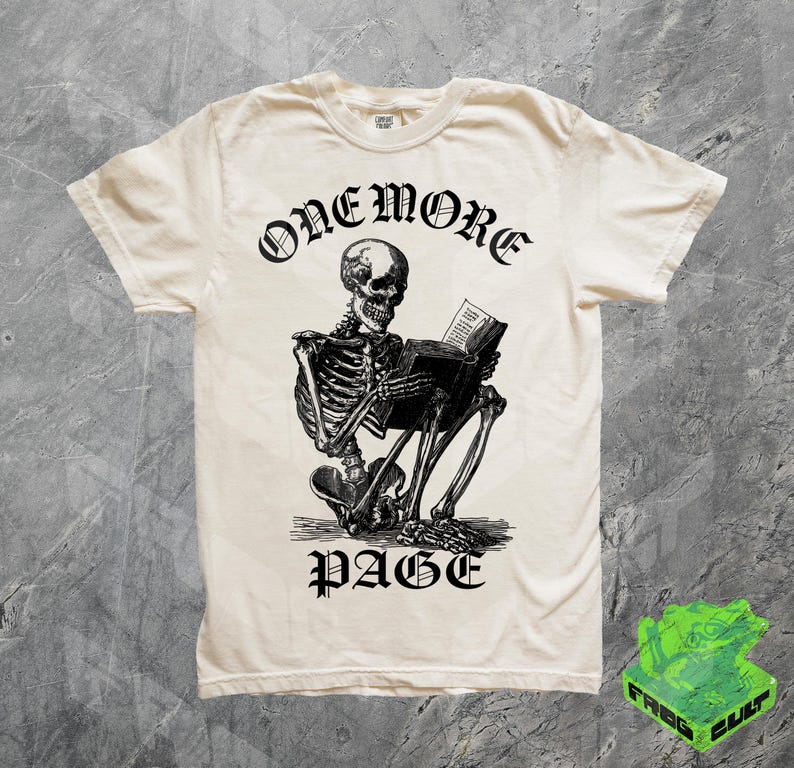 May include: Cream-colored t-shirt featuring a black illustration of a skeleton reading a book. The text "ONEMORE" is arched above the skeleton, and "PAGE" is below. The shirt has a classic crew neck and short sleeves.