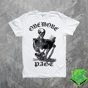 May include: White t-shirt featuring a black and white illustration of a skeleton reading a book. The text "ONE MORE PAGE" is arched above the skeleton, with "PAGE" below. The shirt has a crew neck and short sleeves.