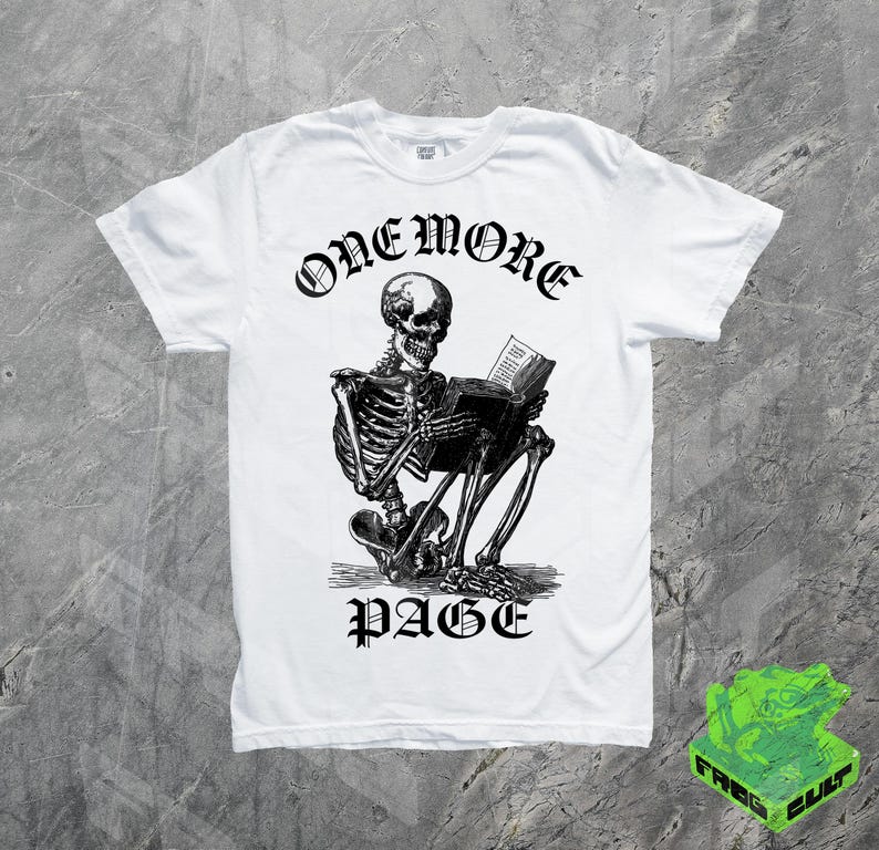 May include: White t-shirt featuring a black and white illustration of a skeleton reading a book. The text "ONE MORE PAGE" is arched above the skeleton, with "PAGE" below. The shirt has a crew neck and short sleeves.