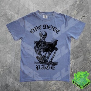 May include: A light blue t-shirt featuring a black illustration of a skeleton reading a book. The text "ONEMORE PAGE" is arched above the skeleton. The shirt has short sleeves and a crew neck.