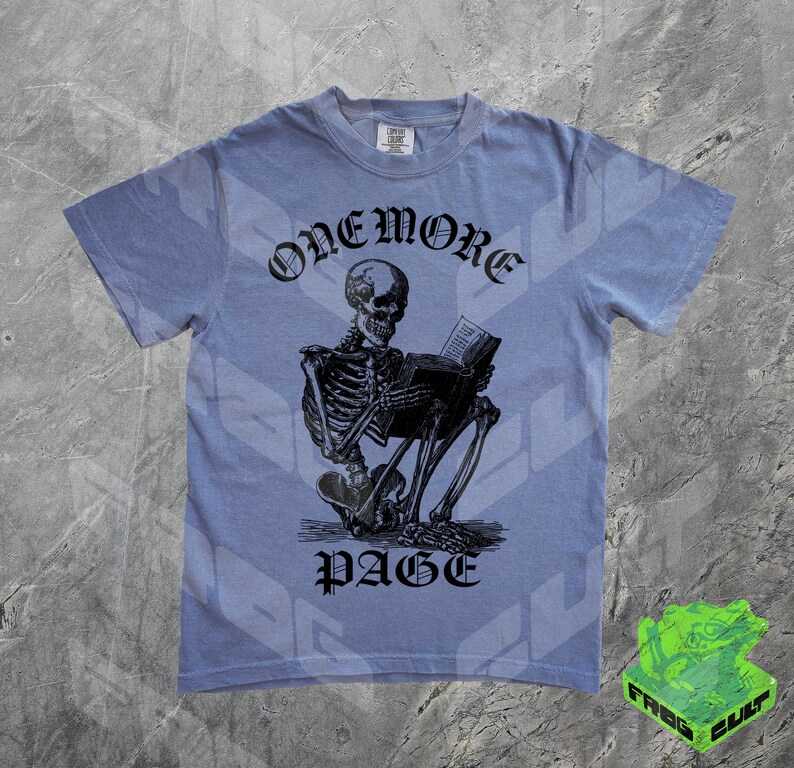 May include: A light blue t-shirt featuring a black illustration of a skeleton reading a book. The text "ONEMORE PAGE" is arched above the skeleton. The shirt has short sleeves and a crew neck.