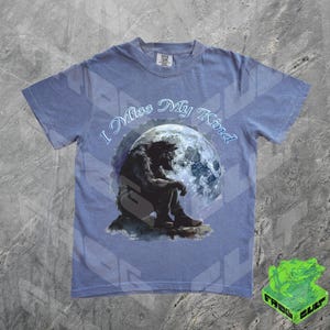 May include: A light blue t-shirt with a graphic of a werewolf sitting under a full moon. The text "I Miss My Kind" is printed on the shirt.