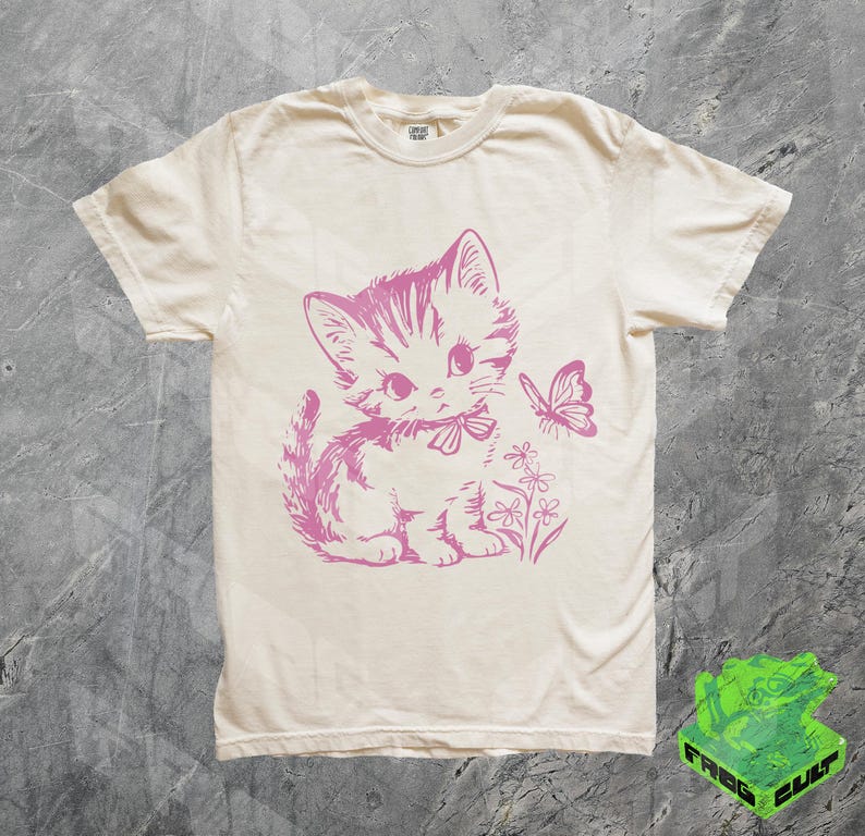 May include: Cream-colored t-shirt featuring a pink illustration of a kitten with a bow, a butterfly, and flowers. The shirt has short sleeves and a crew neck. The text "CONVERT FASHION" is printed on the collar.