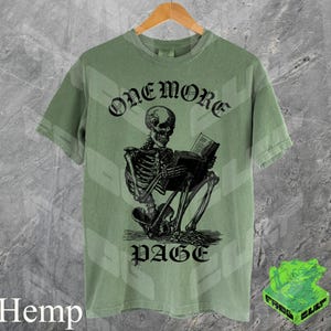 May include: Sage green t-shirt with a black graphic of a skeleton reading a book. The text "ONEMORE PAGE" is above and below the skeleton. The shirt is hanging on a wooden hanger.