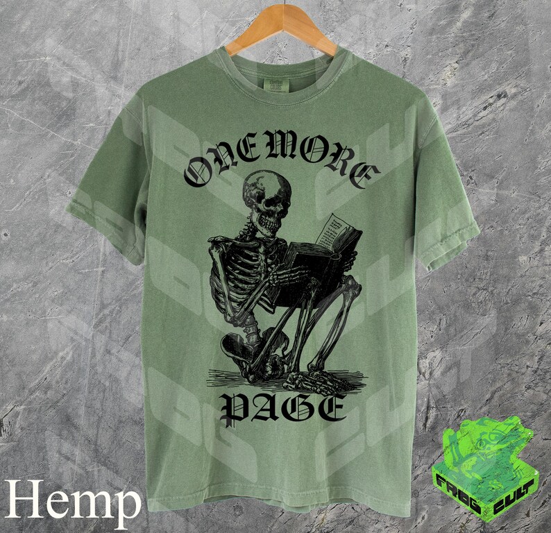 May include: Sage green t-shirt with a black graphic of a skeleton reading a book. The text "ONEMORE PAGE" is above and below the skeleton. The shirt is hanging on a wooden hanger.