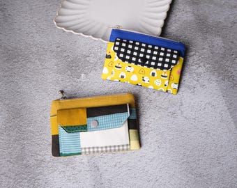 May include: Two handmade fabric pouches. One is yellow with a black and white checkered flap and a blue zipper. The other is mustard yellow with a patchwork design of various fabrics, including blue, green, and white. A white plate is in the background.