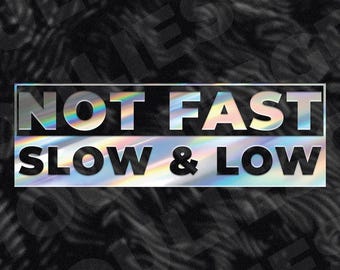 May include: A rectangular sticker with the text "NOT FAST" above "SLOW & LOW". The text is in a holographic, iridescent font, set against a black and gray patterned background. The sticker has a silver border.