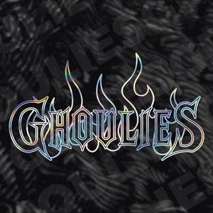 May include: A holographic sticker with the word "GHOULIES" in a stylized font, outlined in iridescent colors. The design includes flame-like accents above the text, set against a black and gray swirling background.