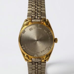 May include: Close-up of the back of a gold-toned wristwatch with a silver-toned metal band. The watch face is silver with gold-toned accents. The back of the watch has text that reads "WATER RESISTANT" and "ONE METAL 3035".