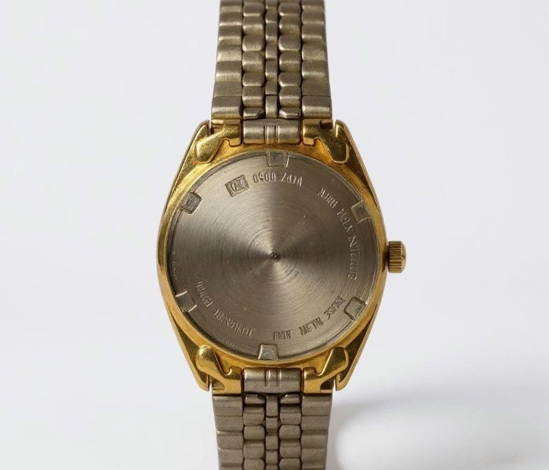 May include: Close-up of the back of a gold-toned wristwatch with a silver-toned metal band. The watch face is silver with gold-toned accents. The back of the watch has text that reads "WATER RESISTANT" and "ONE METAL 3035".
