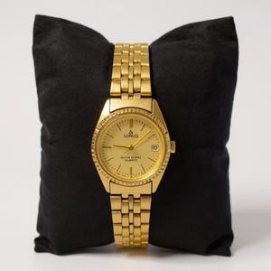 May include: A gold-toned Lorus quartz watch with a matching link band, displayed on a black pillow. The watch face is gold with black hands and markers, and a date window at the 3 o'clock position. The watch is labeled "WATER RESIST QUARTZ".