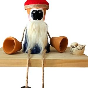 May include: A whimsical gnome figurine made from terracotta flower pots. The gnome is wearing a red hat, a blue shirt, and a white beard. The gnome is sitting on a wooden shelf with a small bird figurine beside it.