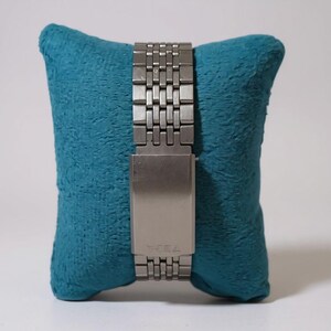 May include: A silver-toned watch band with a rectangular clasp is displayed on a teal blue pillow. The watch band is made of small, rectangular metal links. The clasp has the word "TEOA" engraved on it.