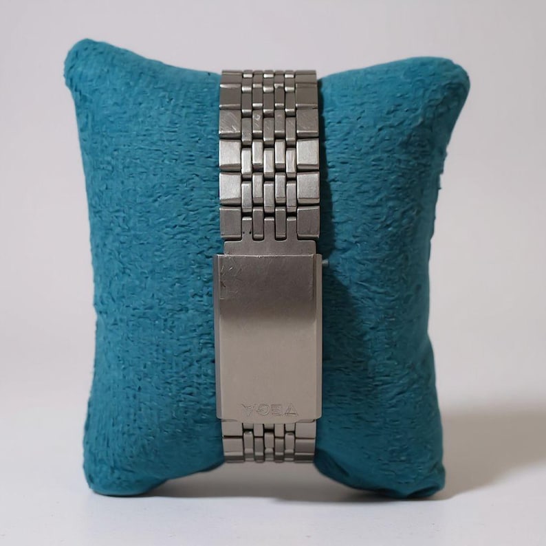 May include: A silver-toned watch band with a rectangular clasp is displayed on a teal blue pillow. The watch band is made of small, rectangular metal links. The clasp has the word "TEOA" engraved on it.