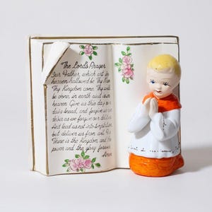 May include: A ceramic figurine of a child in prayer, with a book-shaped base. The book displays "The Lord's Prayer" with floral accents. The child figure wears a white robe with an orange sash and yellow hair.