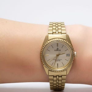May include: A gold-toned Lorus wristwatch with a round face and a textured bezel. The watch has a gold band and a date display at the 3 o'clock position. The watch face is light-colored with black hour markers and hands.