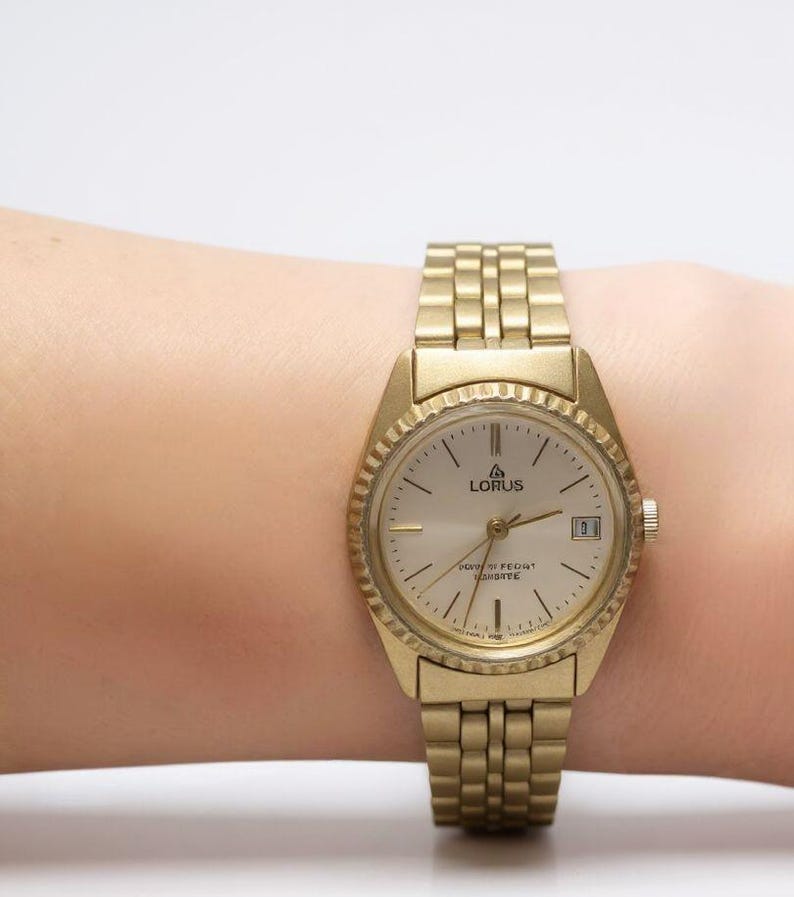 May include: A gold-toned Lorus wristwatch with a round face and a textured bezel. The watch has a gold band and a date display at the 3 o'clock position. The watch face is light-colored with black hour markers and hands.