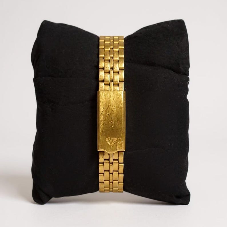 May include: A gold-toned watch with a rectangular clasp and a linked band, resting on a black pillow. The clasp features a small, stylized design. The watch band is made of small, rectangular links.