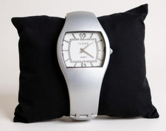 May include: Silver wristwatch with a white face and black numerals, resting on a black pillow. The watch face has the word "TERGOV" and "QUARTZ" printed on it. The watch band is a continuous silver band.