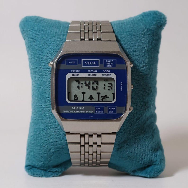 May include: A silver-toned digital watch with a blue face, displaying the time as 7:40:13 AM. The watch, branded "VEGA", features multiple function buttons and a metal band. It rests on a teal blue cushion.