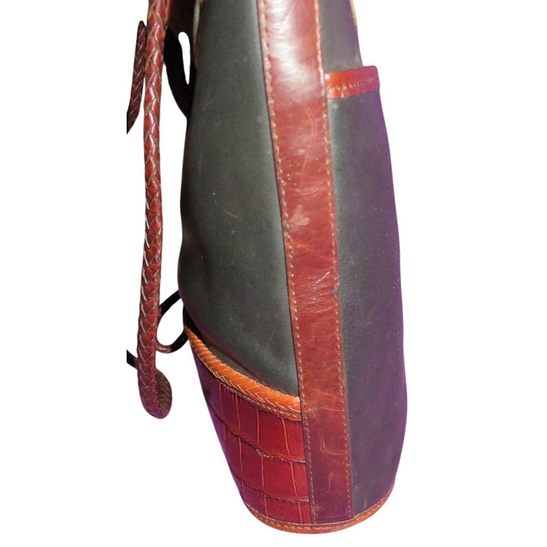 May include: A black and brown handbag with a braided strap. The bag features a dark brown leather trim and a textured, reddish-brown bottom panel. The bag has a side pocket.