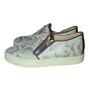 May include: A pair of slip-on sneakers with a snakeskin pattern in shades of gray and beige. The shoes feature a brown zipper detail with a gold-colored zipper pull and a white sole. A gold-colored emblem is visible on the side.