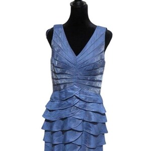 May include: A sleeveless, blue dress with a V-neck and layered, ruffled skirt. The bodice features horizontal pleats, and the dress is displayed on a black mannequin. The fabric has a subtle sheen, giving it a formal appearance.
