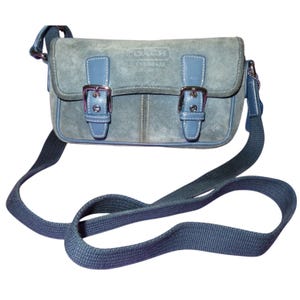 May include: A blue suede crossbody bag with a long adjustable strap. The bag features a flap closure with two silver buckles and light blue leather accents. The Coach logo is visible on the front.