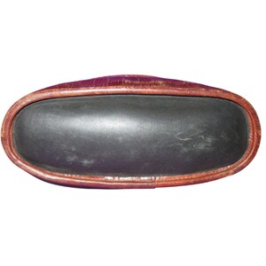 May include: An oval-shaped, dark gray leather tray with a brown leather trim. The tray has a slightly raised edge and a smooth, matte finish. The leather appears to be aged, with some visible wear and tear.